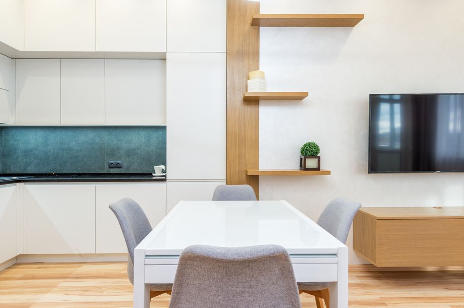 Modern apartment interior with table with chairs next to shelves and open kitchen with cabinets near TV on wall